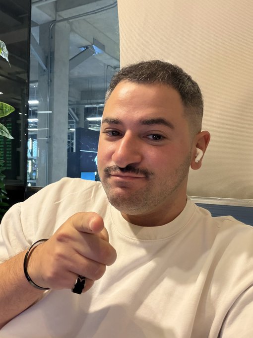 A man with short dark hair and a light beard wearing a white t-shirt points at the camera with AirPods in his ears, smiling in what appears to be an office setting.