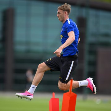 A soccer player in a blue training jersey and black shorts runs around orange training cones on a grass field.