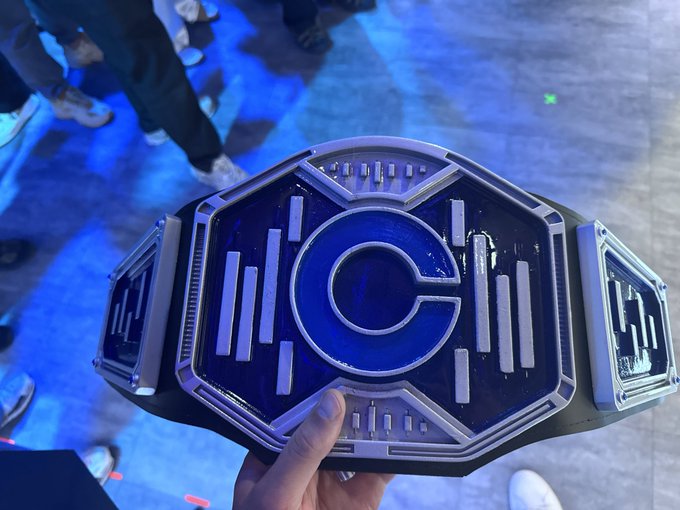 A close-up of an ornate, futuristic championship wrestling belt with a large silver 'C' centerpiece on a blue background, being held up at what appears to be an event.