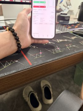 A person wearing a beaded bracelet holds a smartphone displaying what appears to be a trading app, seated at a desk with a chart-patterned mousepad, with slippers visible below.