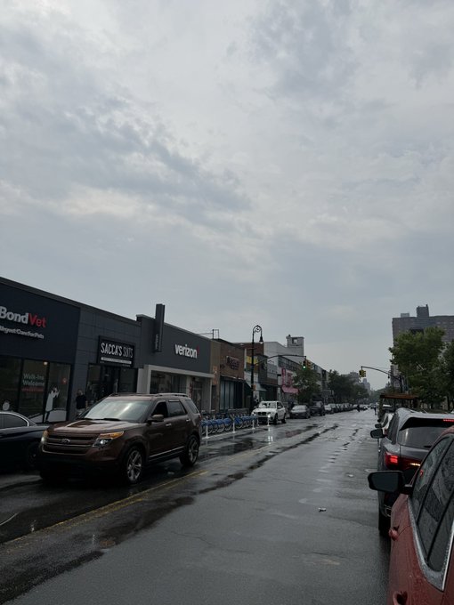 A wet city street under a cloudy sky features storefronts including Bond Vet, Sacks, and Verizon, with cars parked along the road.