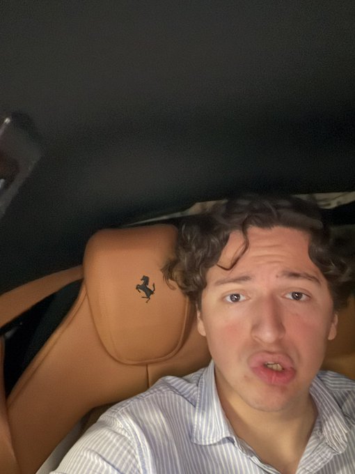 The image shows a man sitting inside a car, wearing a striped shirt with his head resting against the back of the seat. The car has a tan interior, and there's a black logo visible on the passenger side door handle. The man appears to be looking at the camera or something else in front of him, possibly posing for a photo.