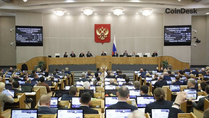 A wide shot of the Russian State Duma parliamentary chamber with the Russian coat of arms and flag at the front, watermarked with the 'CoinDesk' logo in the upper right.