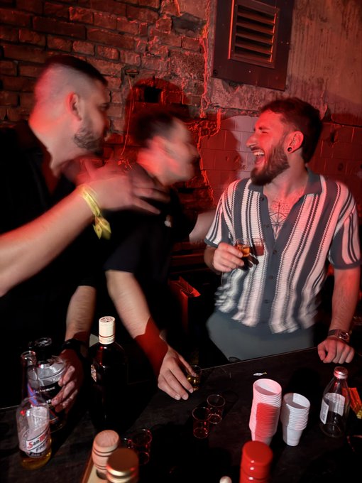 Several men gather at a dimly lit bar with red lighting, one with a mohawk appears blurred in motion while another in a striped shirt laughs and holds a drink.