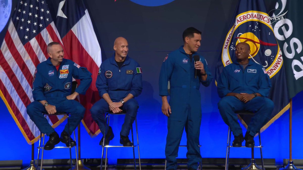 The image features four men dressed in blue suits, sitting on chairs with their legs crossed. They are positioned behind a stage that has an American flag and a NASA flag displayed above them. The men appear to be engaged in conversation or listening to a speech, possibly related to space exploration or the moon.