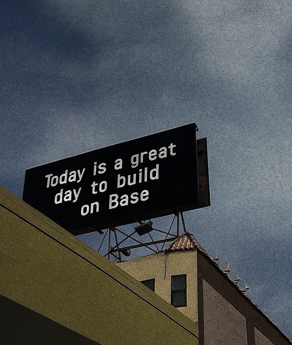 A billboard against a cloudy sky reads "Today is a great day to build on Base," reinforcing the post's optimistic message about developing on the Base platform. The sign is mounted above a building with a tiled roof, photographed from a low angle.