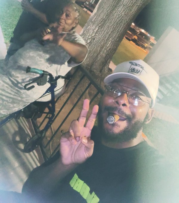 A man wearing a white 'Phillies' cap and glasses holds a peace sign while smoking a cigar, with a woman posing behind him near a bicycle and tree at night.