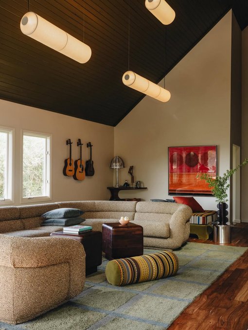 A cozy living room with a vaulted dark wood ceiling, cylindrical pendant lights, guitars hanging on the wall, a large beige sectional sofa, and abstract red artwork.