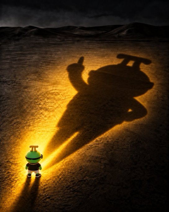 A small cartoonish figure with a green helmet stands in a barren landscape, casting a large, imposing shadow of itself with a thumbs-up gesture under a dramatic sunset.