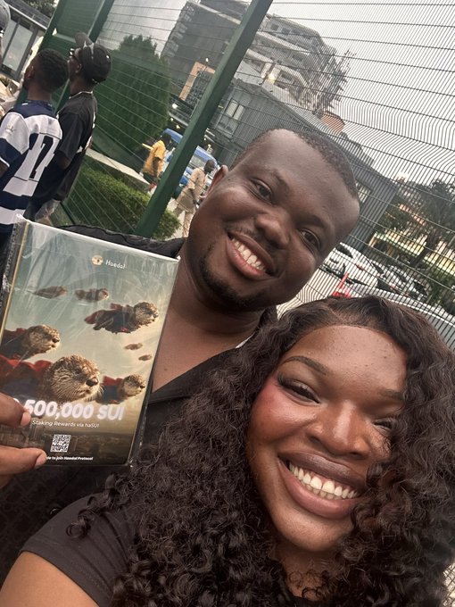 A vertical selfie of a smiling man and a woman, with the man holding up a 'Handel' SupInfog package containing '500,000 SUI' tokens, taken outdoors in front of a green fence.