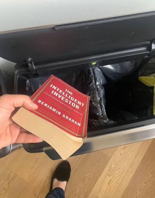 A person discards a copy of "The Intelligent Investor" by Benjamin Graham into a trash bin, symbolically rejecting time-tested value investing principles in favor of the post's promotion of risky memecoin speculation.