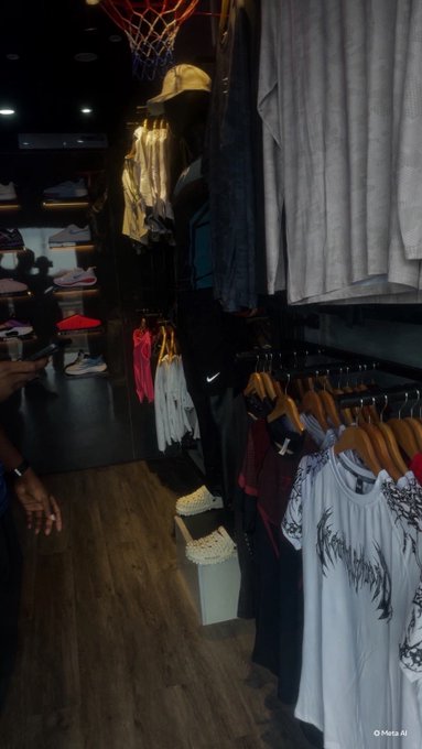 The interior of a clothing store shows racks of apparel including shirts and hoodies, with sneakers displayed on shelves and a basketball hoop hanging from the ceiling.