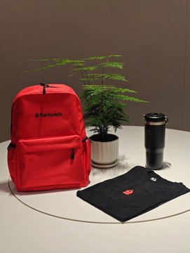 A red Backpack-branded backpack, a black travel tumbler, and a black t-shirt with red and white logos are displayed on a white table next to a potted plant, showcasing the giveaway prizes being offered in partnership with Backpack.