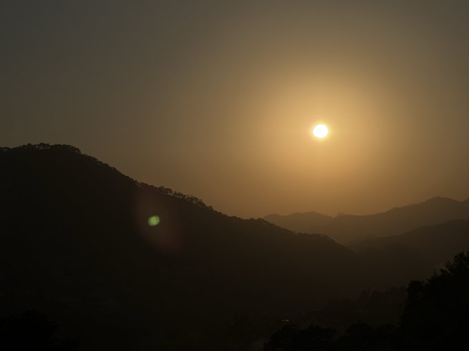The image shows a beautiful sunset over a mountain range with trees visible in the background, creating a serene atmosphere. The sun is setting behind the mountains, casting a warm glow across the scene. The sky appears hazy, adding depth and contrast to the landscape.