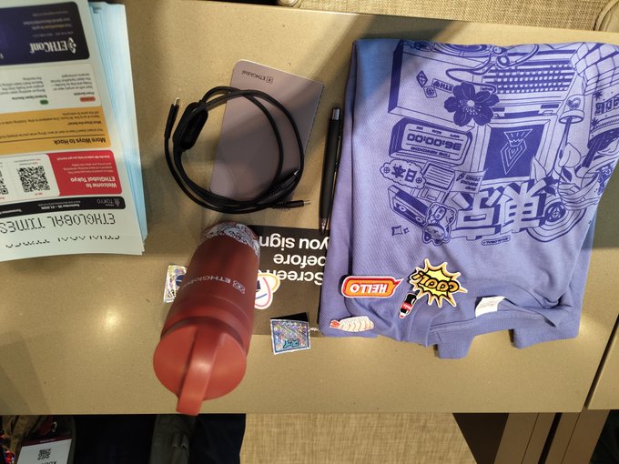 An overhead shot of event swag on a table, including a 'NEXON' branded lanyard, a black power bank, a pen, a red water bottle, and a blue graphic t-shirt with Japanese-style artwork and stickers.