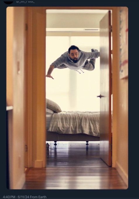 A man in a hoodie appears to be floating horizontally in mid-air above a bed in a bedroom, captured in what looks like a social media post from Earth.