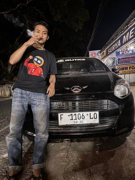 A man stands beside a Daihatsu car with license plate "F 1106 LO" while smoking, with a "SARI BUMI ME" sign visible in the background, fitting the post's mention of smoking in the evening.