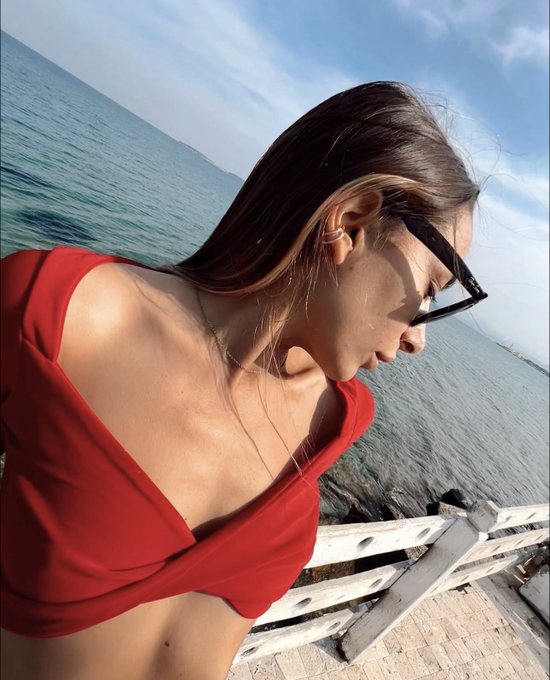 A woman with long brown hair, wearing a red top and sunglasses, looks down while standing near a coastal railing with the sea in the background.