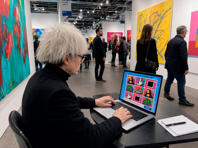 An elderly woman with white hair works on a laptop displaying pop art images in what appears to be an art fair, with colorful paintings on the walls and other attendees in the background.
