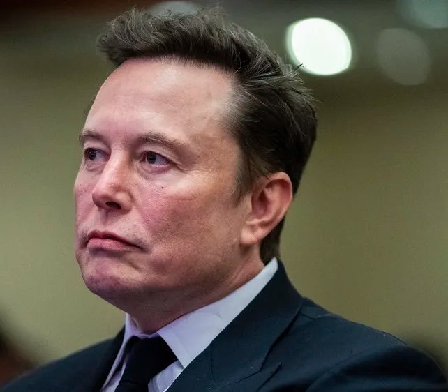 Elon Musk, wearing a dark suit and tie, gazes off-camera with a serious expression in this close-up portrait, the image accompanying a political update where he urges voters to hold senators accountable over their stances on the SAVE America Act and stalled CLARITY Act.