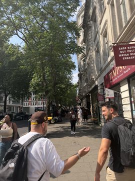 Two men wearing backpacks and caps walk down a sunny city sidewalk near a restaurant with a 'THIS REPUBLICAN CITY' sign visible on the building.