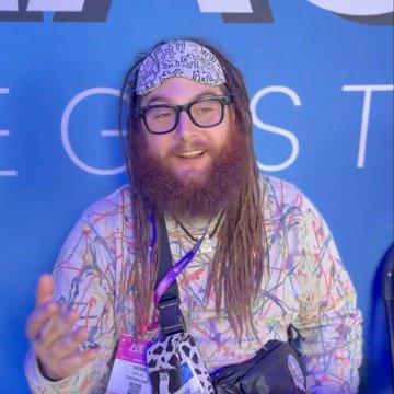 A bearded man with glasses, a patterned kippah, and long sidelocks, wearing a colorful patterned shirt and a lanyard, smiling in front of a blue backdrop.