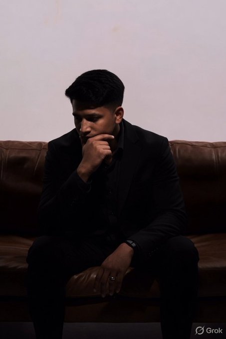 A young man in a dark suit sits pensively on a brown leather couch with his hand to his chin, in a dimly lit room with a watermark reading 'Grok' in the bottom right corner.