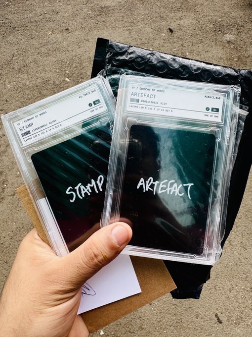 A hand holds two CGC-graded Economy of Words cards—"Stamp" and "Artefact"—each displayed in protective slabs over a black padded mailer, illustrating Jack Butcher's minimalist conceptual art series.