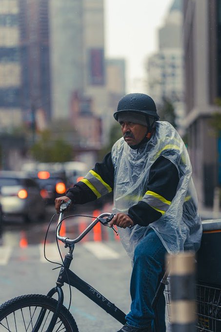 A man wearing a black helmet and a clear plastic rain poncho over a dark jacket rides a bicycle down a wet city street lined with tall buildings.