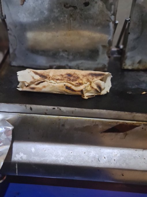 The image features a sandwich sitting on top of an industrial grill, likely used for cooking food like burritos or wraps. The sandwich appears to be partially burnt, indicating that it has been cooked recently. In the background, there are two more sandwiches visible, one placed closer to the left side and another towards the right side of the image.