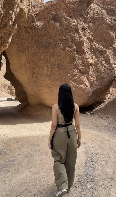 The image shows a woman walking away from the camera while wearing tan pants and a brown tank top. She appears to be enjoying her time outdoors as she walks along a dirt path or trail, with a large rock formation visible behind her. The photo is titled "gm to ordinals, runes, bitmaps, rare sats, brc20 and bitcoin enjoyers".
