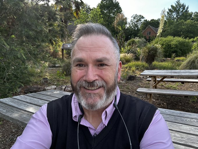 A smiling older man with gray hair and a beard, wearing a black sweater vest over a light purple shirt, taking a selfie outdoors in a garden setting with picnic tables and greenery behind him.
