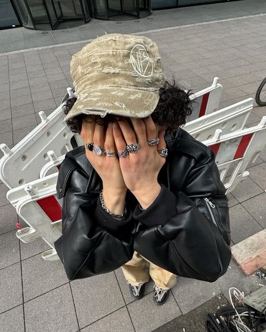 A person wearing a black leather jacket and a distressed olive-green cap sits behind a white road barrier, covering their face with ring-covered hands.