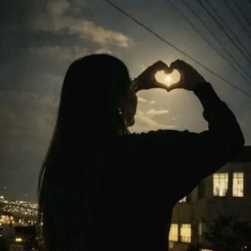 A silhouette of a person with long hair forming a heart shape with their hands against the sun, with city lights visible in the background.