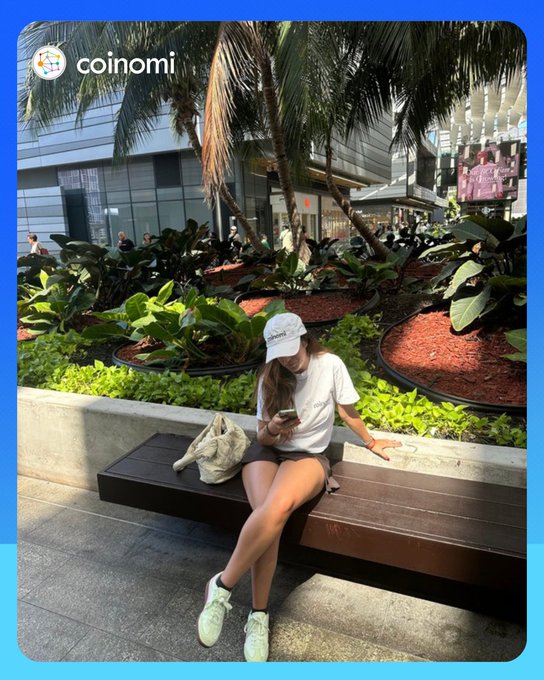 A promotional image for Coinomi wallet showing a young woman in a white cap and t-shirt sitting on a bench looking at her phone, set in a landscaped urban area with palm trees and planters.