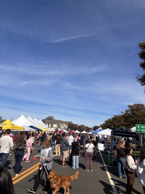 A crowd of people walks through a street fair or market under a bright blue sky, lined with white and yellow tents on both sides, with a golden retriever walking in the foreground.