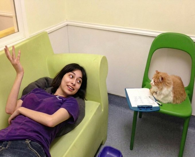 A young woman in a purple shirt lies on a green couch, smiling and gesturing, while a fluffy orange cat sits on a nearby green chair with a clipboard, appearing to conduct an interview.