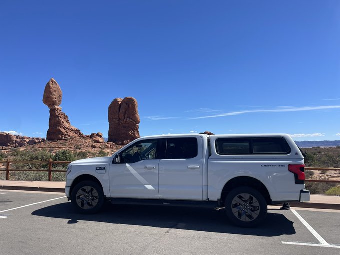 A side profile of a white Ford F-150 Lightning Limited pickup truck parked in a lot, with the iconic red rock formations of Arches National Park visible in the background.