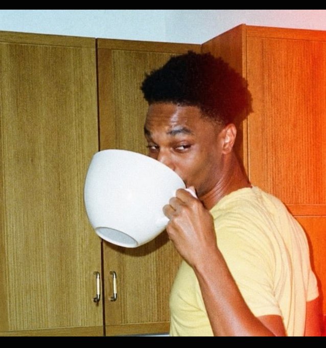 A young man in a yellow t-shirt drinks from an oversized white bowl while looking directly at the camera, standing in a kitchen with wooden cabinets.
