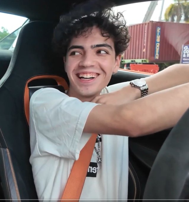 A young man with curly dark hair wearing a white t-shirt and silver chain sits in the driver's seat of a car with an orange seatbelt, smiling broadly while gesturing with his arms.