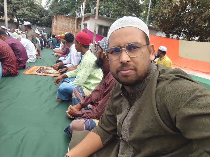The image shows a man wearing glasses sitting down, possibly at an event or gathering with other people around him. The man has a green shirt on and appears to be posing for a photo. In the background, there are several other people who seem to be engaged in conversation or observing something happening nearby. The post includes text that reads "Good morning my X family Eid mubarak All friends @NomismaNetwork 3look Bullish @useTria @sleepagotchi @River4fun".