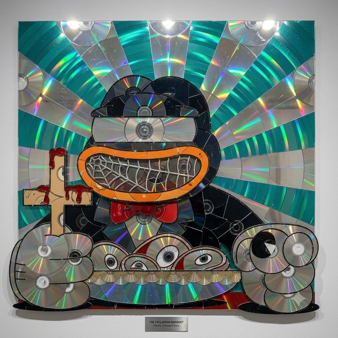 A photograph of an art installation titled 'THE CYCLOPEAN BANQUET' by Moore, Compact Discs, made from repurposed CDs and DVDs forming a creature-like figure with a radiant background.