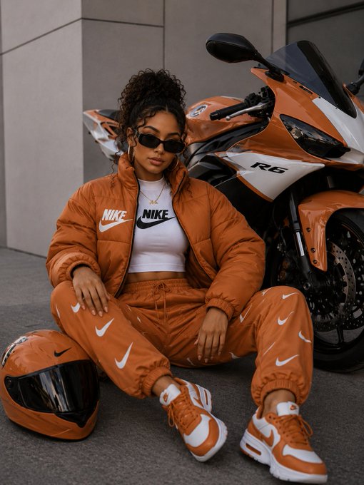 The image features a woman sitting next to an orange and white motorcycle, wearing a Nike jacket and matching shoes. She has her legs crossed while posing for the photo. The background of the image shows a building with a gray facade, which complements the overall color scheme of the scene. The post caption reads "gm fam", indicating that this is likely a sponsored content piece featuring the woman and the motorcycle.