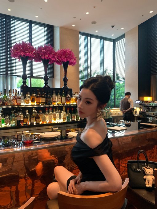 A young woman in a black off-shoulder dress sitting at a bar, with shelves of liquor bottles, pink flowers, and a bartender in the background.