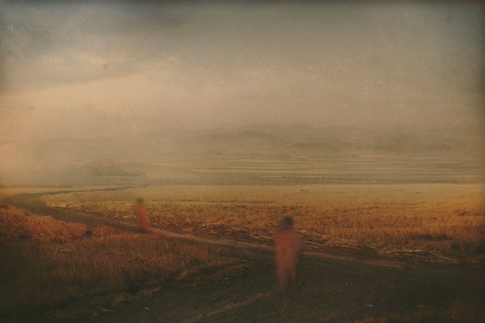 A hazy, vintage-style photograph shows two blurred figures walking along a dirt path through a vast, foggy landscape with mountains in the distance.