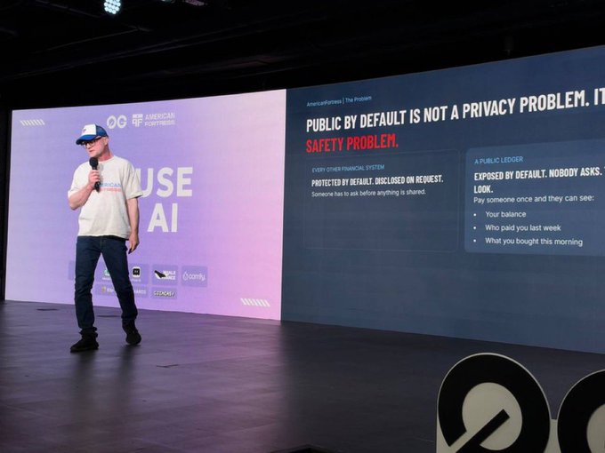 A man in a white cap and t-shirt speaks on stage next to a large screen displaying 'PUBLIC BY DEFAULT IS NOT A PRIVACY PROBLEM. IT'S A SAFETY PROBLEM.'