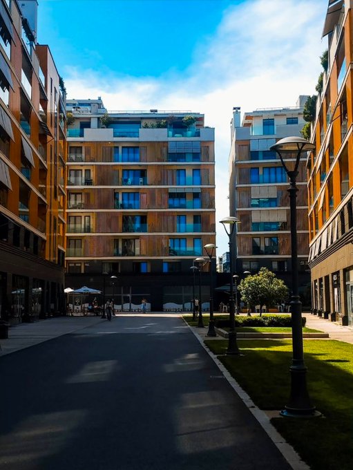A modern urban courtyard features multi-story residential buildings with wood-paneled facades, decorative lamp posts, and a paved walkway under a blue sky.