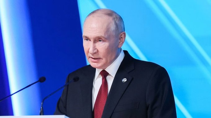 A man in a black suit and red tie speaks at a podium with microphones, set against a blue background.