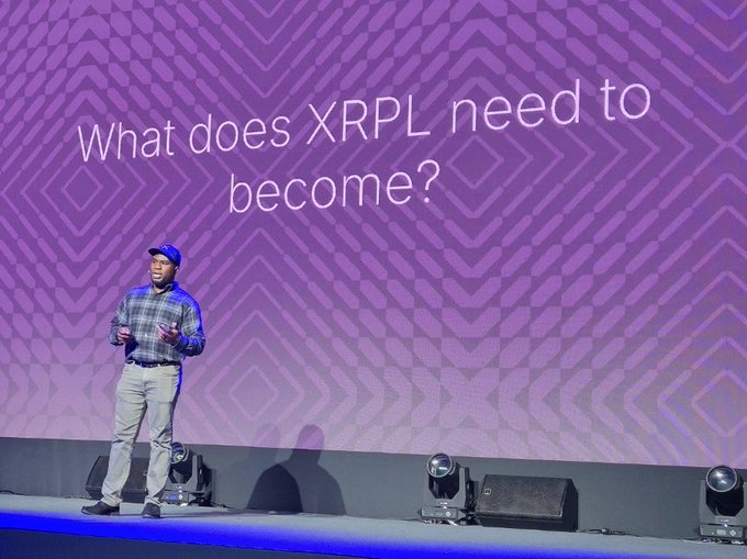 A man in a plaid shirt and cap speaks on stage at XRP Seoul 2026, with the slide behind him posing the question "What does XRPL need to become?"—setting the thematic stage for Ripple's presentation on the ledger's evolution toward confidential transfers, transaction batching, and AI agent wallets.