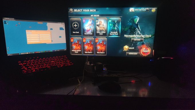 A photograph of a gaming setup in a dark room featuring two monitors, with the main screen displaying a deck selection screen for the card game 'Mythic' showing options like 'New Deck', 'Starter Deck', and a 'Fight' button.